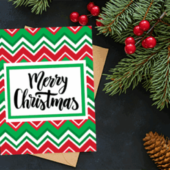 A Printable Christmas Card featuring red, green, and white zigzag patterns with "Merry Christmas" sits on a dark surface beside evergreen branches, red berries, and a pine cone.
